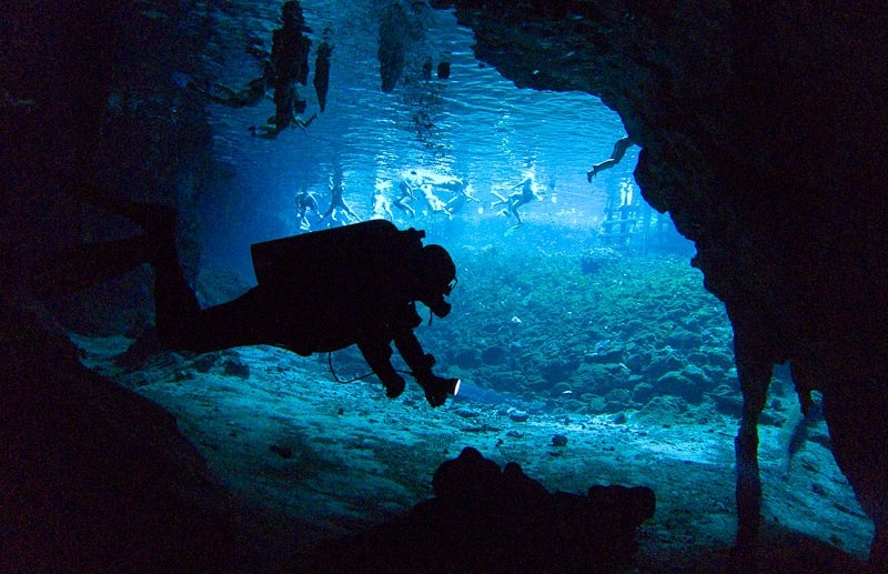 Un buceador explora las partes más inaccesibles del gran Cenote de Sac Actun, con un grupo de turistas al fondo.