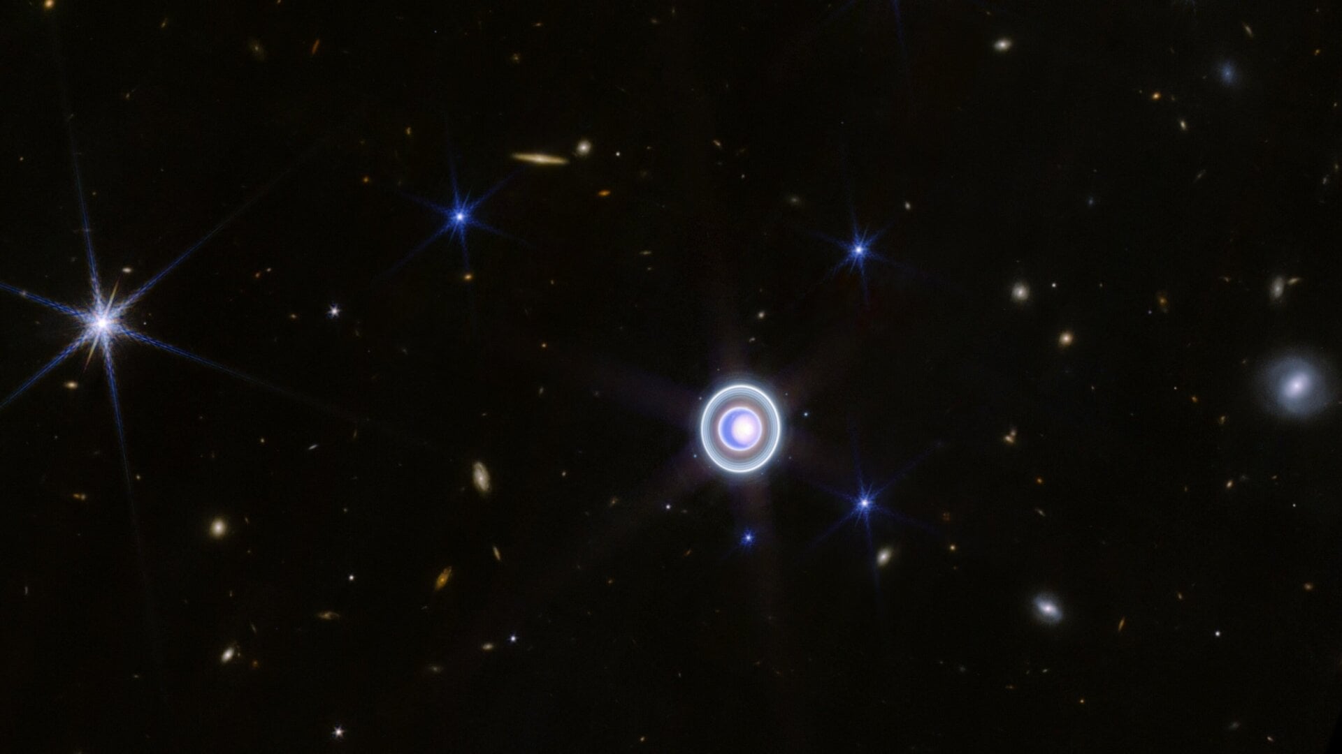 Urano y sus anillos, rodeados por un puñado de galaxias y estrellas de fondo.